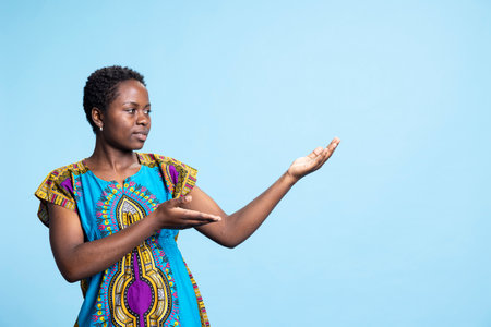 African american female model creating promotional web ad on camera, pointing at left or right sides for social media marketing advertisement. Young adult smiling in studio, cultural ethnic attire.の写真素材