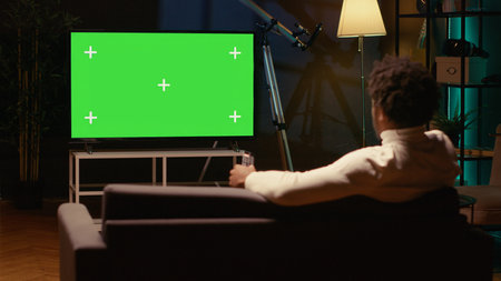 African american man zapping through isolated screen TV channels using remote, relaxing on couch in living room used as home theatre. Person using mockup television set to enjoy leisure timeの写真素材