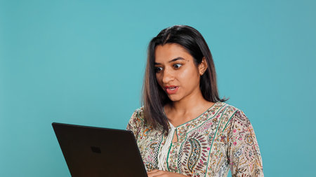Indian woman composing email, typing on laptop, frustrated with malfunctioning keyboard. Person sending mail online, struggling to use defective notebook, studio background, camera Bの写真素材