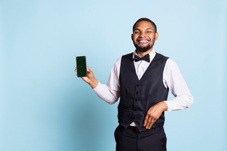 Hotel concierge staff holding a smartphone with chroma key display, showing copy space against blue background in studio. Hotelier bellhop welcoming resort guests at reception.の写真素材