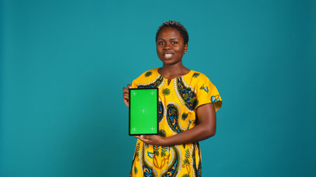 Happy natural girl showing a green screen against blue background, presenting isolated mockup display in studio. Confident young adult in traditional attire with tribal native elements. Camera B.の写真素材