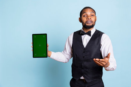 Hotel porter with suit and tie showing green screen on tablet in studio, working in hospitality industry and tourism. Lodging resort manager offers concierge services for guests.の写真素材