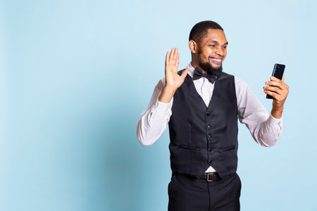 Classy bellboy hotel porter answering videocall on smartphone, discussing with clients or friends against blue background. Doorkeeper on online videoconference connection in studio.の写真素材