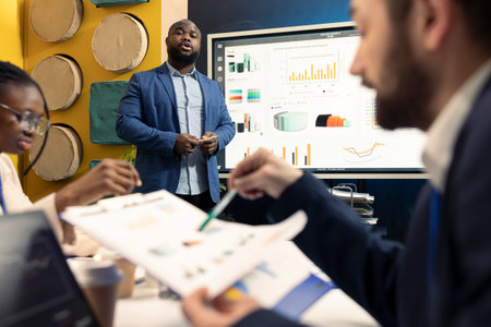 African american office manager providing important information, presenting infographics and details to his staff members before starting a new task, business meeting in boardroom.の写真素材
