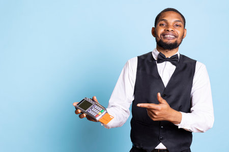 Young receptionist pointing at the pos terminal with credit card in it, waiting for the electronic transaction to pay for lodging at holiday resort. Porter with suit and tie in studio.の写真素材