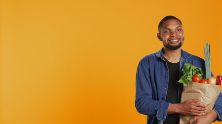 Portrait of smiling model carrying bag full of fresh bio groceries, buying locally grown organic fruits and vegetables from eco friendly supermarket. Vegan guy supports zero waste. Camera B.の写真素材