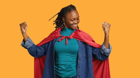 Happy superhero flexing muscles to prove force, listening music, isolated over studio background. Jolly young girl with headphones posing as hero in costume, kissing biceps, showing strength, camera Aの写真素材