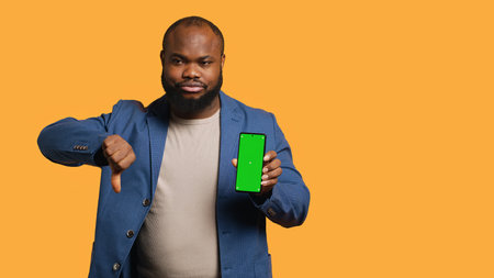 Upset african american man showing thumbs down sign gesturing holding green screen phone,...