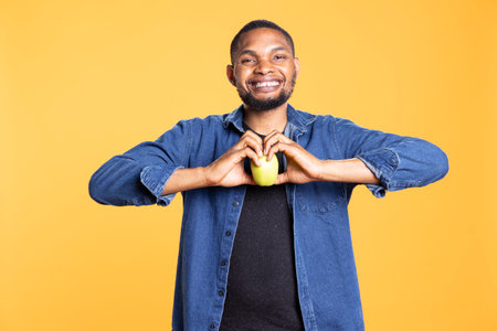 African american smiling guy does heart shape sign with an apple inside of it, advertising healthy...