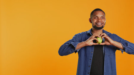 African american relaxed guy does a heart shape sign with an apple, recommending locally grown produce in studio. Happy pleased person enjoys healthy eating and chemicals free nutrition. Camera B.の写真素材