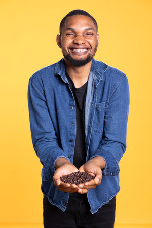 African american cheerful person posing with black roasted coffee beans against yellow background,...