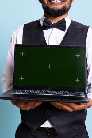 Doorkeeper bellboy holding a laptop with green screen in studio, presenting isolated chroma key display while he works in hospitality industry at a five star hotel. Professional staff.の写真素材