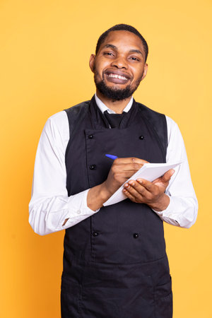 Professional caterer writing clients orders on notepad for the bill, takes notes of desired food against yellow background. Waiter with apron, notebook service for customer satisfaction.の写真素材