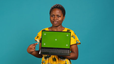 Young adult showing a laptop with isolated mockup screen in studio, presenting a display with green screen against blue background. Female model posing with chroma key. Camera A.の写真素材