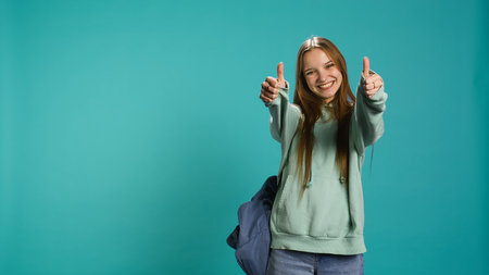 Happy girl doing thumbs up hand gesturing, showing approval, having positive mood. Smiling teenager presenting confirmation, doing agreement finger sign, studio background, camera Bの写真素材