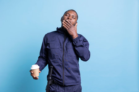 Sleepy african american bodyguard yawning in front of camera, drinks a cup of coffee to get over exhaustion with a caffeine refreshment. Security agent experiences burnout from night shifts.の写真素材