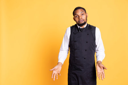 Young butler shrugging and doing i dont know gesture in studio, expressing his uncertainty with a doubtful expression. Restaurant staff waiter in apron acting confused and clueless.の写真素材