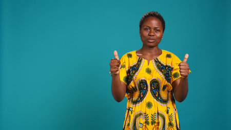 Cheerful positive woman showing thumbs up gesture in studio, presenting her approval with a like sign against blue background. Young smiling person shows satisfaction. Camera B.の写真素材