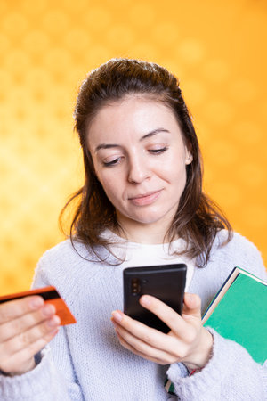 Smiling woman holding books, purchasing more with phone, conveying joy of reading concept, studio background. Cheerful person buying novels online, showing appreciation for literatureの写真素材