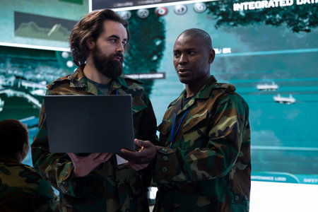 Diverse team of IT operators comparing real time battlefield data from a laptop to a big screen,...