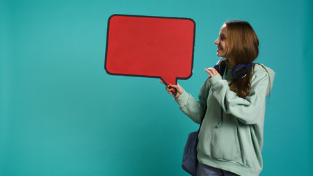 Smiling woman holding red speech bubble sign of empty copy space for message. Joyous teenager presenting thought bubble cardboard used as promotion concept, isolated over studio background, camera Bの写真素材