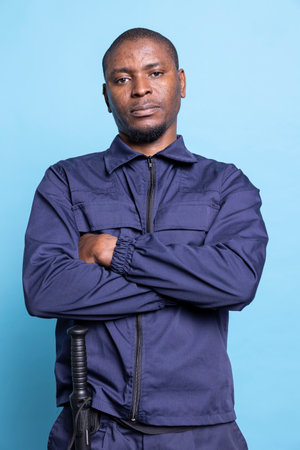 Portrait of african american guard posing with arms crossed against blue background, wearing his professional uniform with confidence and pride. Serious armed agent works on patrol service.の写真素材