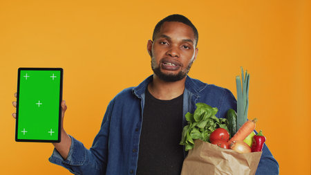 Vegan adult holding a tablet with greenscreen layout in studio, carrying paper bag full of ethically sourced chemicals free produce. Guy advocating for healthy eating with mockup screen. Camera A.の写真素材