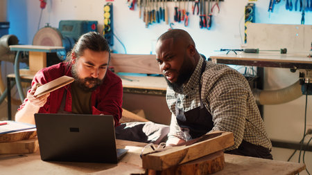 Carpenter and colleague verifying finished wood art product, comparing with blueprint. Artisan and apprentice inspecting wooden object for damages based on schematics on laptop screen, camera Aの写真素材