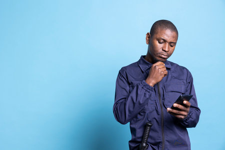 African american security guard navigating online browser on phone, texting messages on his work break. Agent in uniform checks social media accounts and apps against blue background.の写真素材