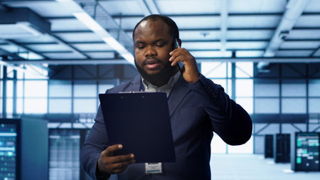 IT professional looking over data center checkup checklist paperwork, talking on phone in server farm. African american man surrounded by server rigs, chatting during telephone call, camera Aの写真素材