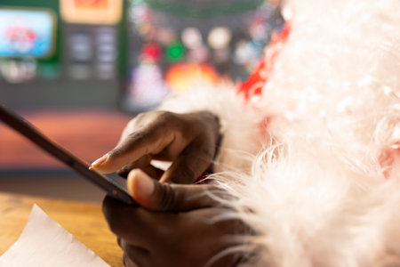 Santa Claus uses a tablet to navigate online webpages in his holiday decorated North Pole log cabin. Enjoying the festive and Christmas traditions, making gift delivery preparations. Close up.の写真素材