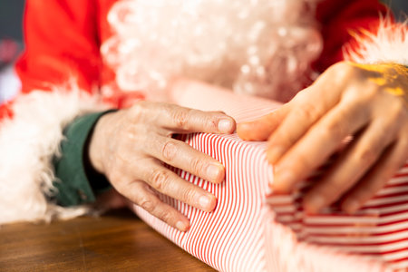 Saint Nicholas wraps a festive gift box surrounded by Christmas lights and ornaments in his workshop. Santa prepares for the holiday season, packing presents to bring joy to children. Close up.の写真素材