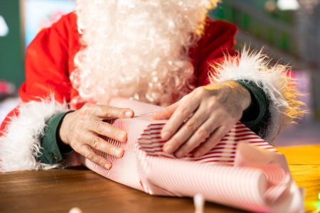 Father Christmas decorates a beautifully wrapped present box with festive wrapping paper, spreading the holiday spirit with lights and ornaments. Santa prepares gifts in his North Pole workshop.の写真素材