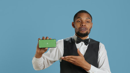 Hotel concierge personnel showing chroma key display on a smartphone, posing with hospitality industry uniform in studio. Doorkeeper lounge area staff shows green screen. Camera A.の写真素材