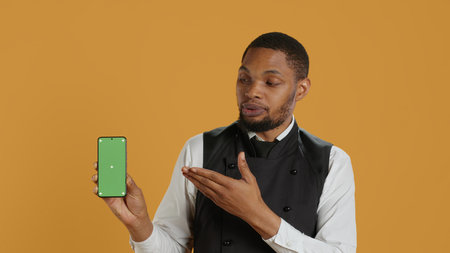Restaurant staff showing a smartphone with green screen in studio, prepared to serve food orders in service oriented environment. Waiter with apron and uniform serving clients at tables. Camera A.の写真素材