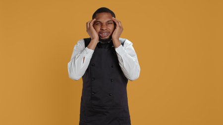 Restaurant employee suffering from a headache and being sick, working too much and feeling unwell with a painful migraine. Waiter in uniform and apron works in hospitality industry. Camera B.の写真素材