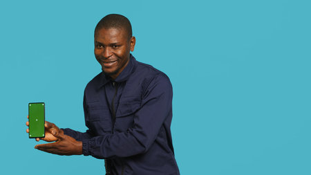 Portrait of security agent presenting mockup phone and securing assigned post, studio background. Guardian showing green screen smartphone while handling security breachesの写真素材