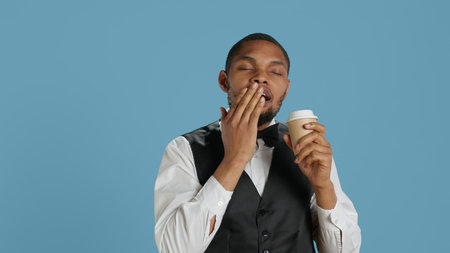Hotel concierge staff drinks coffee while he is yawning and feeling tired, trying to get more energy with the caffeine beverage. Doorkeeper employee is exhausted and fatigued. Camera A.の写真素材