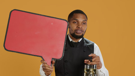 Catering employee showing a red speech bubble icon in studio, carrying a cruet set with condiments. Waiter with professionalism presenting an empty cardboard sign as an advertisement. Camera A.の写真素材