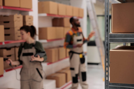 Selective focus of shelves full with cardboard boxes, in background diverse warehouse coworkers preparing clients orders. Storage room employees managing parcels transportation, working in storehouseの写真素材