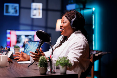 Cheerful black female talk show host engaging with her online audience on live broadcast. African American woman with wireless headset, sitting in home studio recording a podcast episode.の写真素材