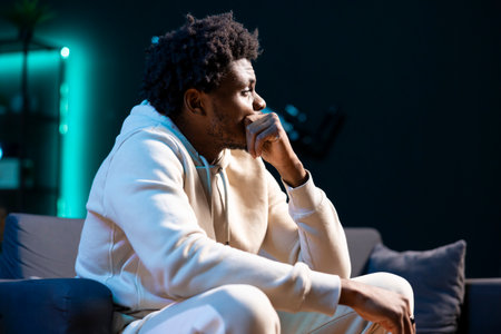 African american man sitting at home on couch late at night, overthinking about problems. Person...
