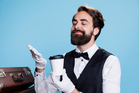 Stylish doorman enjoying smell of coffee over blue background, feeling happy while he holds with cup of drink on camera. Classy skilled hotel porter smells refreshment savour.の写真素材