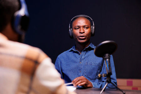 Black guy guest speaks into microphone, addressing questions from radio program host. African American man is interviewed while utilizing audio equipment to record an online chat show at home.の写真素材