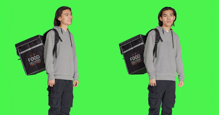 Asian guy works on food delivery with thermal backpack, standing over studio greenscreen backdrop....