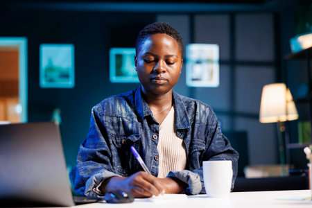Young black woman studying while using her laptop and taking notes on a notepad. African American female entrepreneur browsing the internet and writing down new business ideas.の写真素材