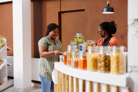 African american client at cashier desk purchasing homegrown organic produce at local food market. Cheerful black female customer assisted by friendly vendor at checkout counter in bio grocery shop.の写真素材
