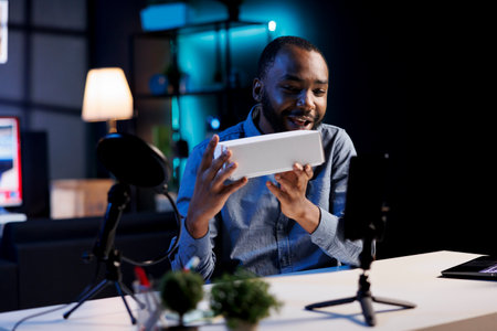Smiling tech content creator in dark studio filming technology review of newly launched digital device, unboxing it and presenting specifications to audience. Media star showing product to viewersの写真素材