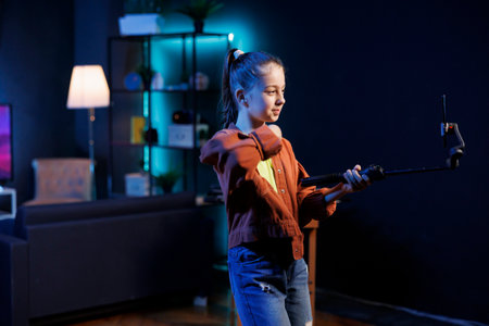 Kid in dimly lit studio joins viral dance craze, recording family friendly video with cellphone on selfie stick. Little girl in living room does latest internet dancing challenge, films with phoneの写真素材