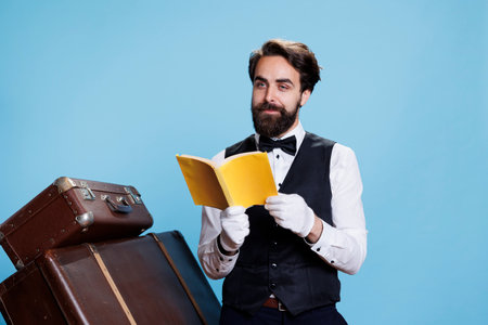 Hotel porter enjoying lecture in studio while he stands next to trolley bags, reading pages of literature or fiction. Classy elegant doorman having fun with stories on camera, seeking knowledge.の写真素材